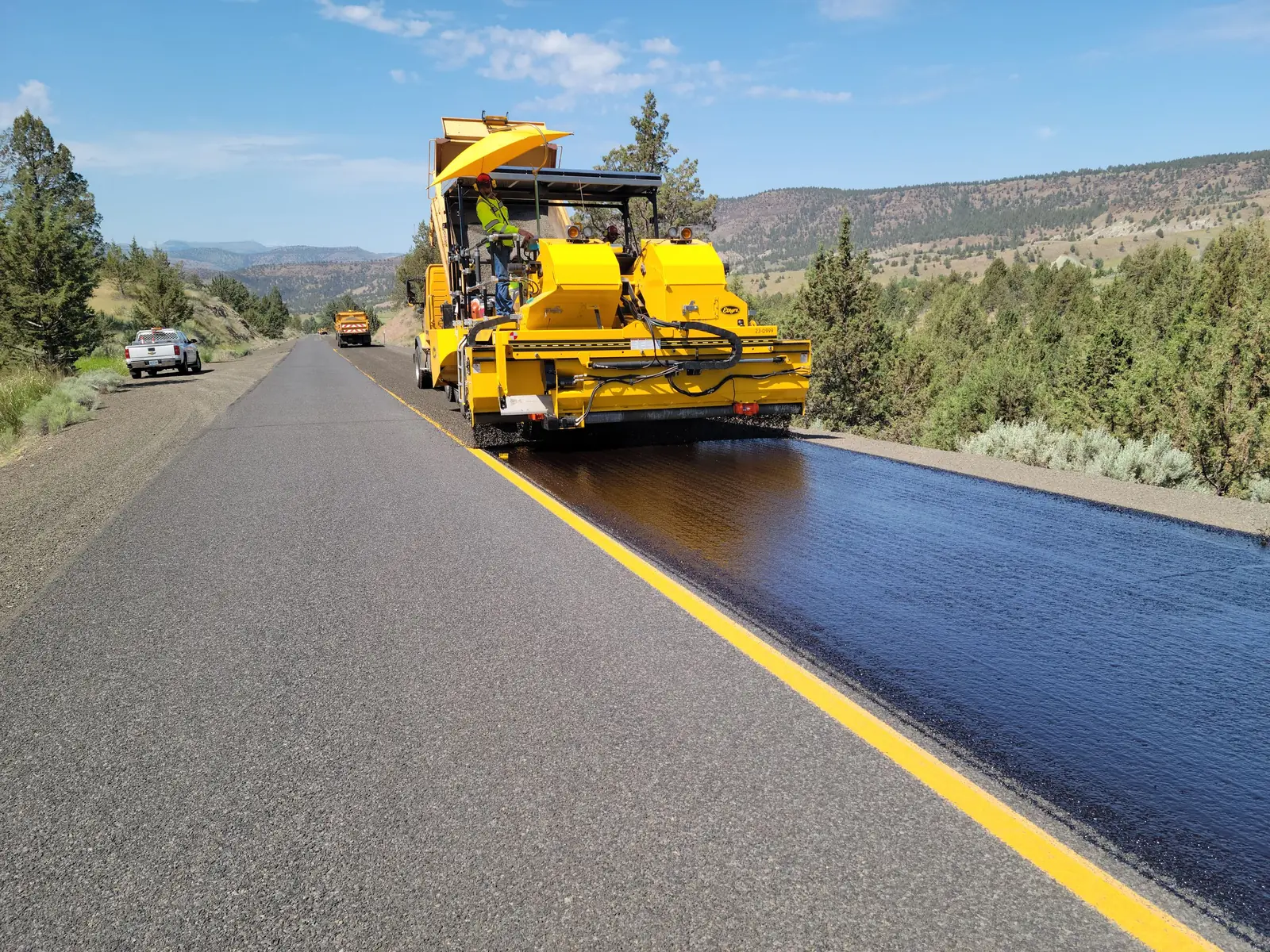 An asphalt paver laying a fresh black mat beside the yellow edge line of a two-lane rural highway.