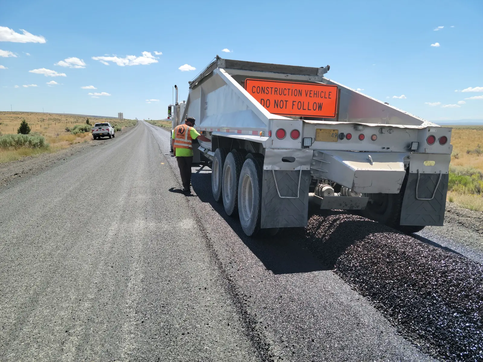 The rear of a haul truck spreading base stone on a highway job, a flagger walking alongside.